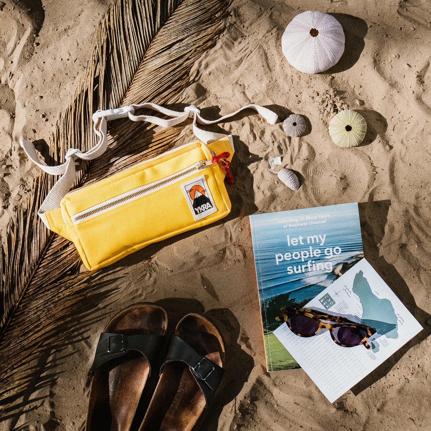 Gelbe Fanny Pack Gürteltasche von YKRA liegt am Strand im Sand neben Büchern und Sandalen