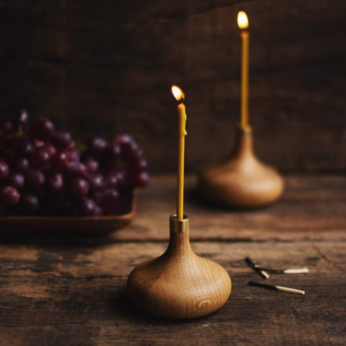 Zwei Kerzenhalter von Ovo Things mit brennenden Ovo Slim Kerzen auf Holztisch stehend im Hintergrund rote Weintrauben