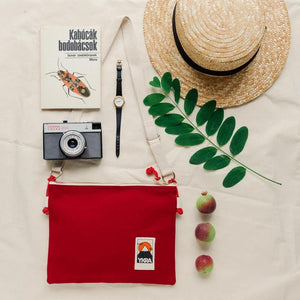 Bordeaux farbende Side Pouch von YKRA neben Sonnenhut und Fotokamera im Retrolook